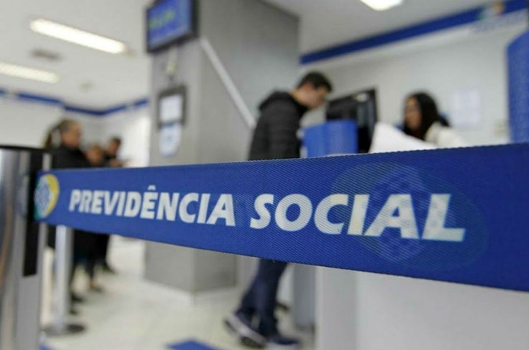 Pessoas aguardam atendimento em agência da Previdência Social com fita azul e letreiro