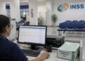 Servidora do INSS analisa requerimentos no computador de uma agência da Previdência Social, com pilha de processos de 2026 sobre a mesa, rotina que fez o volume de análises subir 34,7% e derrubar a fila para 1,6 milhão de pedidos