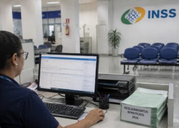 Servidora do INSS analisa requerimentos no computador de uma agência da Previdência Social, com pilha de processos de 2026 sobre a mesa, rotina que fez o volume de análises subir 34,7% e derrubar a fila para 1,6 milhão de pedidos