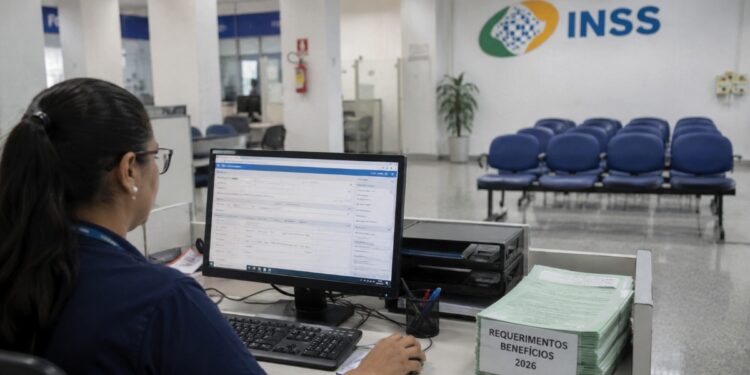 Servidora do INSS analisa requerimentos no computador de uma agência da Previdência Social, com pilha de processos de 2026 sobre a mesa, rotina que fez o volume de análises subir 34,7% e derrubar a fila para 1,6 milhão de pedidos