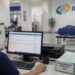 Servidora do INSS analisa requerimentos no computador de uma agência da Previdência Social, com pilha de processos de 2026 sobre a mesa, rotina que fez o volume de análises subir 34,7% e derrubar a fila para 1,6 milhão de pedidos