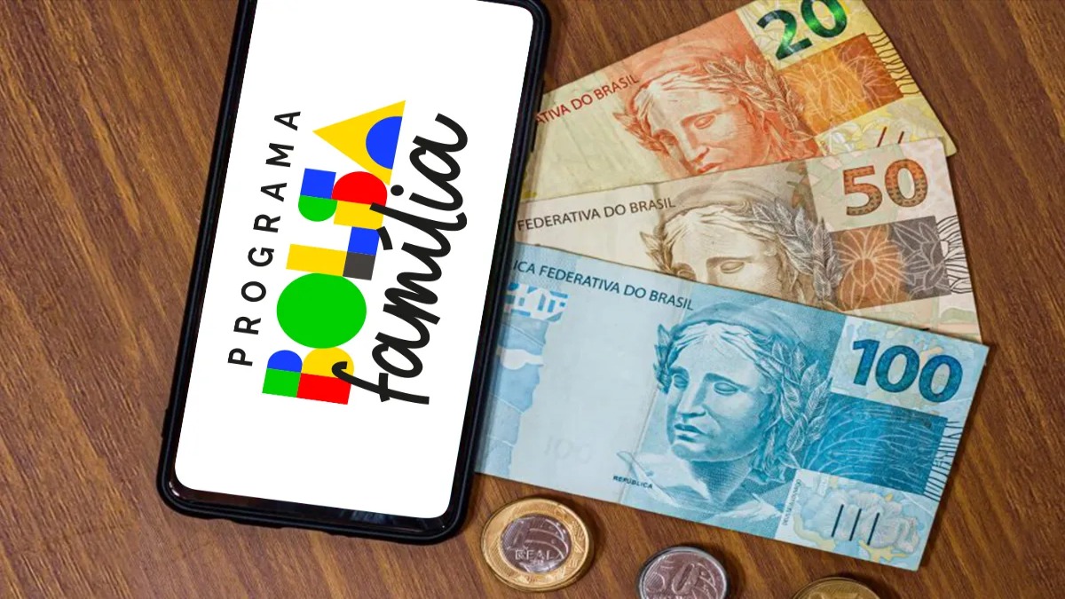 Imagem ilustrativa de celular com a logomarca do Programa Bolsa Família na tela, ao lado de notas de 20, 50 e 100 reais sobre mesa de madeira, cena que representa as quatro formas de usar a parcela de julho de 2026, do aplicativo Caixa Tem ao saque em lotérica