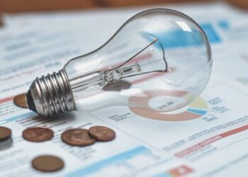 Lâmpada incandescente apoiada sobre faturas de energia elétrica ao lado de algumas moedas