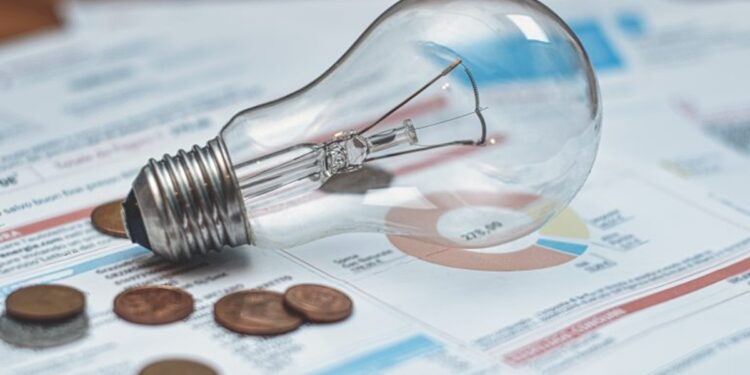 Lâmpada incandescente apoiada sobre faturas de energia elétrica ao lado de algumas moedas