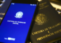 Celular exibindo a tela inicial do aplicativo Carteira de Trabalho Digital apoiado sobre uma carteira de trabalho impressa, em referência ao acesso e à consulta dos contratos