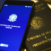 Celular exibindo a tela inicial do aplicativo Carteira de Trabalho Digital apoiado sobre uma carteira de trabalho impressa, em referência ao acesso e à consulta dos contratos