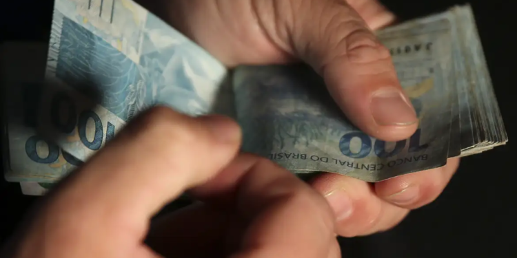 Imagem ilustrativa de mãos contando cédulas de cem reais em um fundo escuro, representando a verba destinada ao pagamento de benefícios sociais