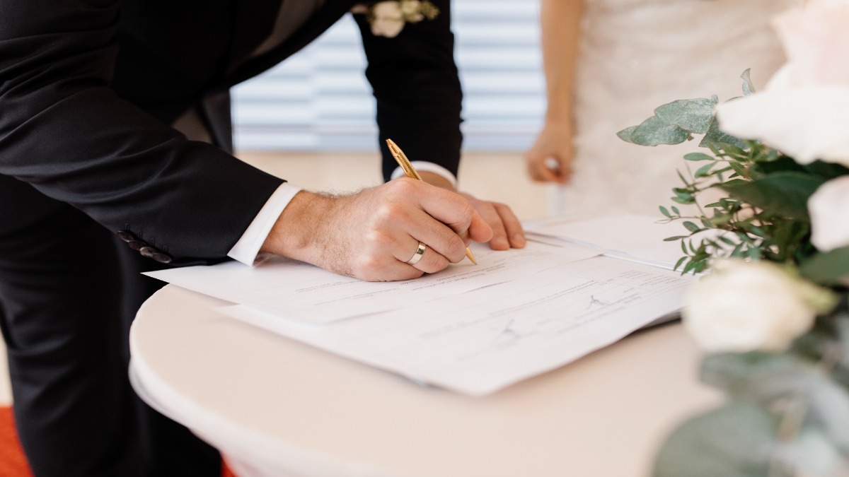 Noivo assina os documentos do casamento sobre uma mesa ao lado do buquê, imagem ilustrativa do registro que gera o livro, a folha e o termo pedidos no formulário da segunda via