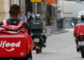 Entregador de aplicativo visto de costas, com mochila térmica vermelha, em rua movimentada onde outros motociclistas trabalham, cena usada para tratar do início do financiamento do Move Brasil