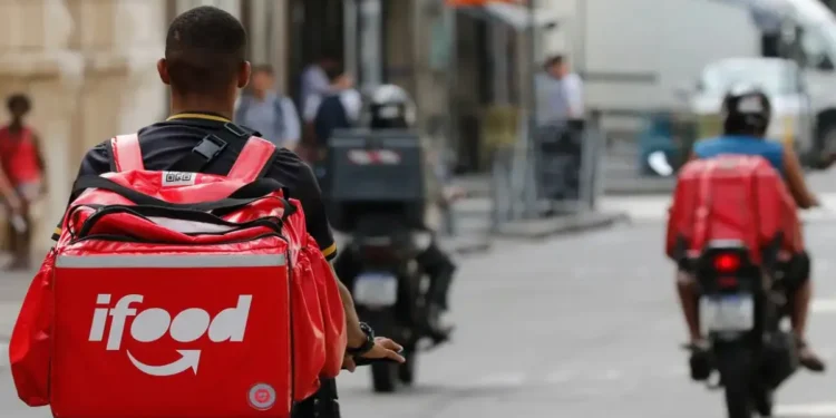 Entregador de aplicativo visto de costas, com mochila térmica vermelha, em rua movimentada onde outros motociclistas trabalham, cena usada para tratar do início do financiamento do Move Brasil