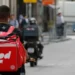 Entregador de aplicativo visto de costas, com mochila térmica vermelha, em rua movimentada onde outros motociclistas trabalham, cena usada para tratar do início do financiamento do Move Brasil