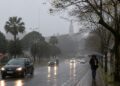 Avenida molhada sob chuva e neblina em cidade do Sul do país, com carros de farol aceso e pedestre de guarda-chuva, cenário previsto pelo INMET com a chegada da frente fria entre 18 e 20 de julho