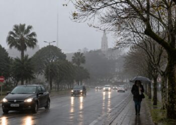 Avenida molhada sob chuva e neblina em cidade do Sul do país, com carros de farol aceso e pedestre de guarda-chuva, cenário previsto pelo INMET com a chegada da frente fria entre 18 e 20 de julho