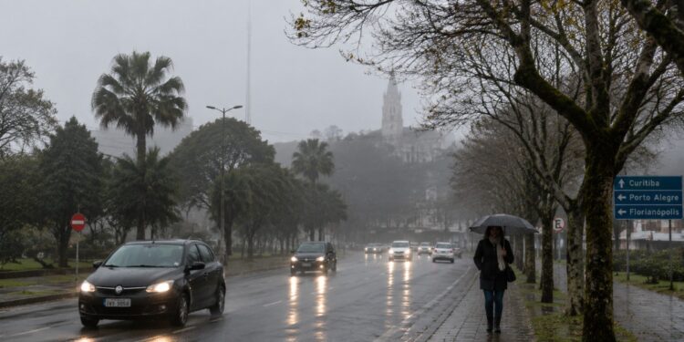 Avenida molhada sob chuva e neblina em cidade do Sul do país, com carros de farol aceso e pedestre de guarda-chuva, cenário previsto pelo INMET com a chegada da frente fria entre 18 e 20 de julho
