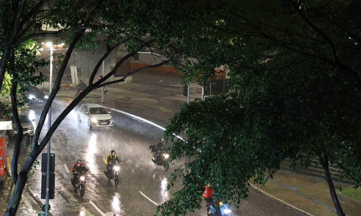 Imagem ilustrativa de chuva forte sobre uma avenida à noite, com motociclistas e um carro trafegando na pista molhada, em referência ao alerta de chuva e ventania