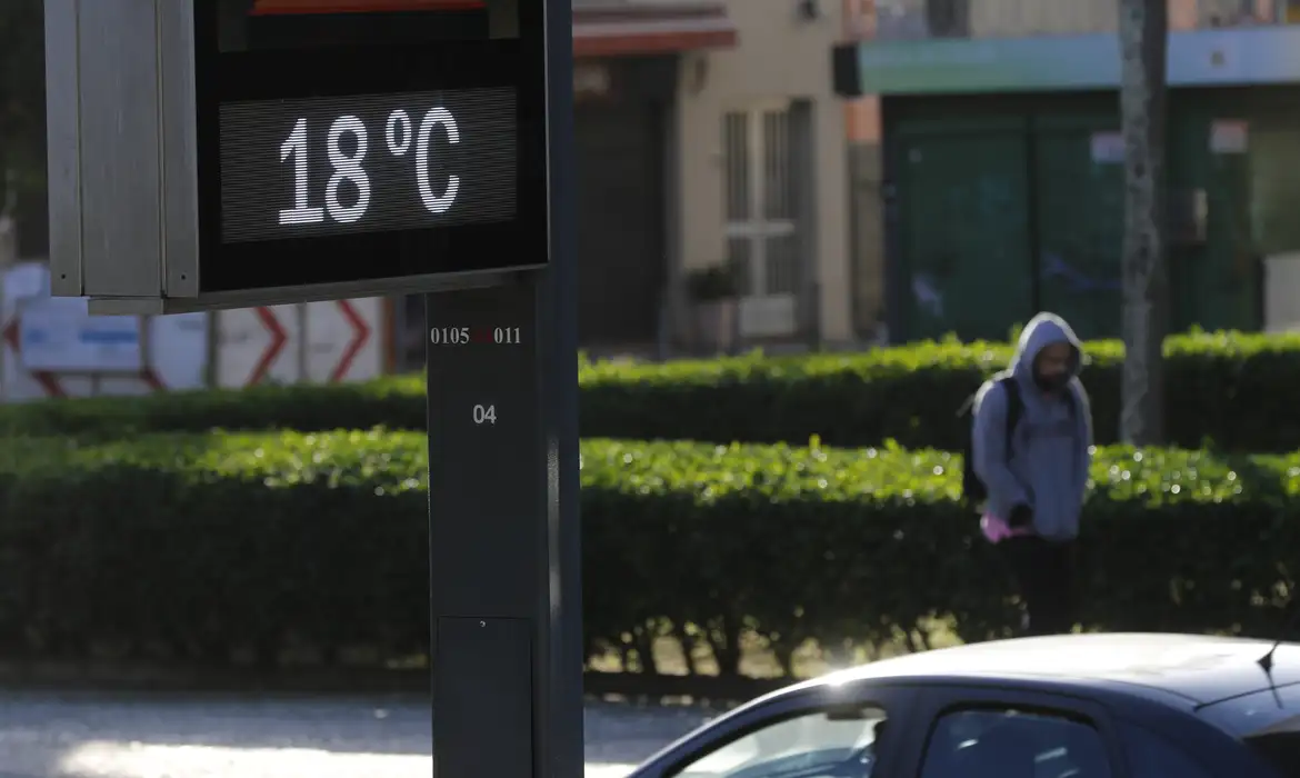Termômetro de rua marca 18 graus e pedestre agasalhado caminha pela calçada, enquanto o INMET aponta chuva no litoral do Nordeste e queda de temperatura com a frente fria no Sul