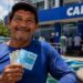 Pescador com notas de R$ 100 em frente a uma agência da Caixa, representando o pagamento do seguro-defeso previsto para esta terça