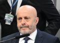 Homem de terno sentado diante de microfone durante depoimento, com crachá do INSS visível ao fundo