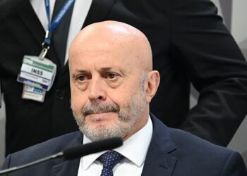 Homem de terno sentado diante de microfone durante depoimento, com crachá do INSS visível ao fundo