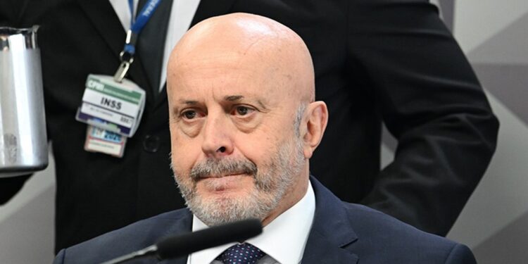 Homem de terno sentado diante de microfone durante depoimento, com crachá do INSS visível ao fundo