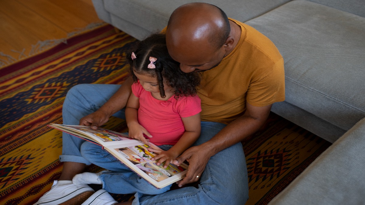10 dias de licença-paternidade a partir de 2027: lei amplia benefício e prevê 20 dias até 2029 2 Pai lê um livro com a filha sentado no chão da sala, convivência ampliada pela nova lei que leva a licença-paternidade de 5 para 20 dias até 2029