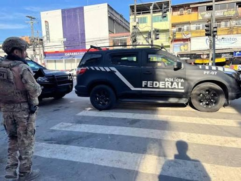 Agente da Polícia Federal ao lado de viatura em cruzamento de rua durante cumprimento de mandados