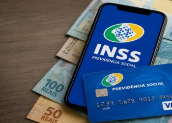 Imagem ilustrativa de celular com a logomarca do INSS na tela, apoiado sobre cédulas de 100 e 50 reais, ao lado de um cartão de benefício da Previdência Social em mesa de madeira, representando o pagamento referente a julho de 2026