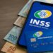 Imagem ilustrativa de celular com a logomarca do INSS na tela, apoiado sobre cédulas de 100 e 50 reais, ao lado de um cartão de benefício da Previdência Social em mesa de madeira, representando o pagamento referente a julho de 2026