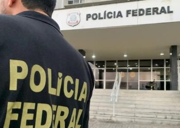 Agente de costas com o colete da Polícia Federal em frente a um prédio da corporação durante operação