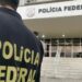 Agente de costas com o colete da Polícia Federal em frente a um prédio da corporação durante operação