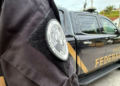 Brasão da Polícia Federal no uniforme de um agente, com viatura estacionada ao fundo