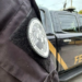 Brasão da Polícia Federal no uniforme de um agente, com viatura estacionada ao fundo