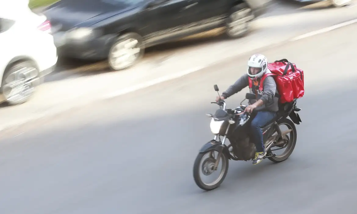 Motociclista de entrega com capacete branco e mochila térmica vermelha trafegando por avenida, cena usada para tratar do cadastro no programa Move Brasil