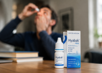 Homem de blusa azul aplica colírio Hyabak sentado em mesa com caixas do produto visíveis