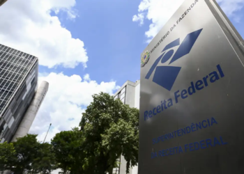 Placa metálica com o logotipo da Receita Federal em frente a prédios da Superintendência, sob céu azul, órgão responsável pela nova regra de desembarque nos aeroportos.