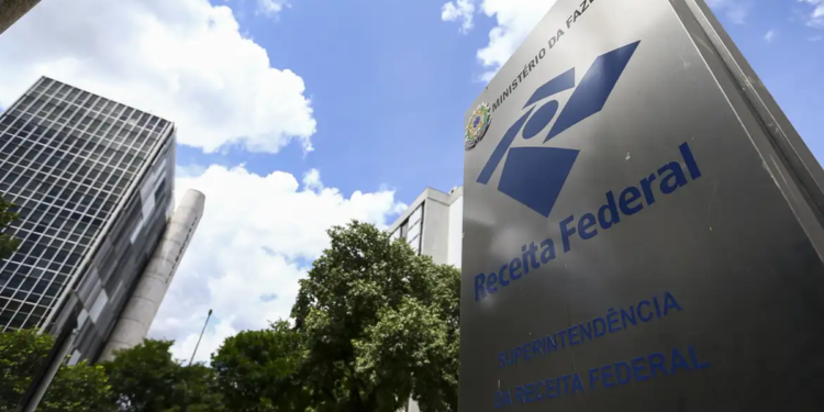 Placa metálica com o logotipo da Receita Federal em frente a prédios da Superintendência, sob céu azul, órgão responsável pela nova regra de desembarque nos aeroportos.
