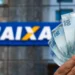 Imagem ilustrativa de mão segurando cédulas de cem reais em leque diante de uma agência da Caixa, representando o pagamento do ressarcimento do PIS/PASEP