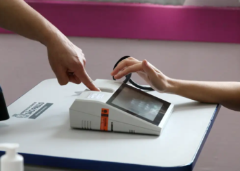 Imagem ilustrativa de eleitor apoiando a mão sobre o leitor biométrico da mesa de votação, enquanto um mesário indica o equipamento, em referência à identificação do eleitorado nas urnas