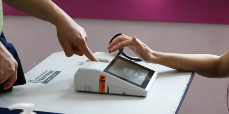 Imagem ilustrativa de eleitor apoiando a mão sobre o leitor biométrico da mesa de votação, enquanto um mesário indica o equipamento, em referência à identificação do eleitorado nas urnas
