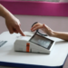Imagem ilustrativa de eleitor apoiando a mão sobre o leitor biométrico da mesa de votação, enquanto um mesário indica o equipamento, em referência à identificação do eleitorado nas urnas