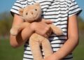Imagem ilustrativa de criança de camiseta listrada segurando com os dois braços um urso de pelúcia, ao ar livre, referência à proteção legal reforçada para crianças e adolescentes