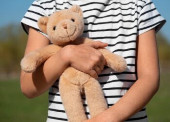 Imagem ilustrativa de criança de camiseta listrada segurando com os dois braços um urso de pelúcia, ao ar livre, referência à proteção legal reforçada para crianças e adolescentes