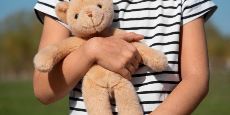 Imagem ilustrativa de criança de camiseta listrada segurando com os dois braços um urso de pelúcia, ao ar livre, referência à proteção legal reforçada para crianças e adolescentes