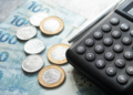 Calculadora preta, caneta, notas de cem reais e moedas sobre mesa.