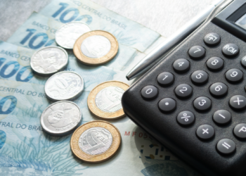 Calculadora preta, caneta, notas de cem reais e moedas sobre mesa.