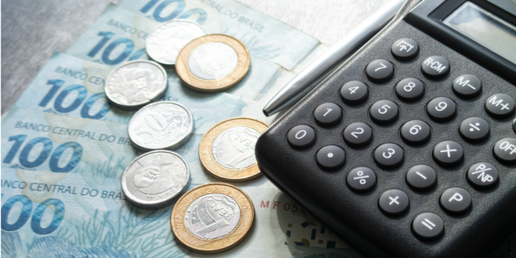 Calculadora preta, caneta, notas de cem reais e moedas sobre mesa.