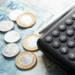 Calculadora preta, caneta, notas de cem reais e moedas sobre mesa.