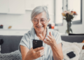 Idosa sorridente celebra boa notícia ao celular em casa.