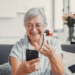 Idosa sorridente celebra boa notícia ao celular em casa.