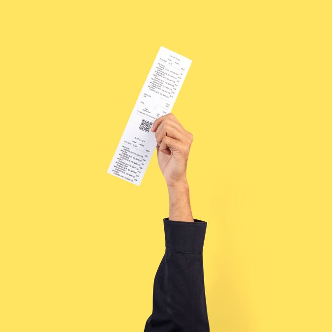 Pessoa exibe nota fiscal detalhada ao alto em fundo amarelo.
